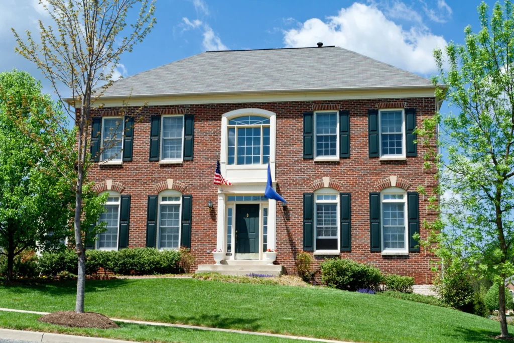 This two-story colonial brick house features a gray asphalt shingle roof, dark green shutters on rows of white-framed windows, and a white-columned entrance that frames a dark front door that pops from afar. 4SeasonsHome in Rockville can elevate your home's curb appeal with these details, no matter what your style.