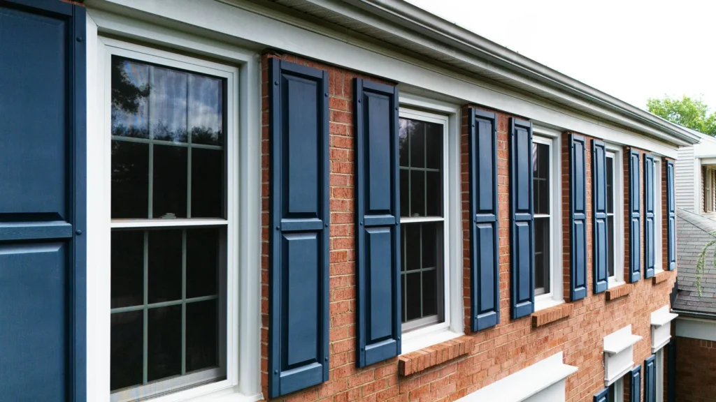 Grey asphalt shingle roof and blue shuttered windows newly installed to a red-brick house surrounded by lush green lawn and tress in the suburbs of Maryland.